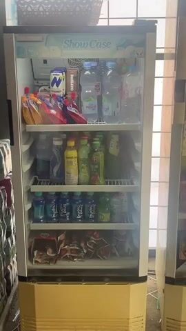 The video shows a commercial display refrigerator filled with various beverages and some packaged snacks. The refrigerator has a glass door and is illuminated from within, making the contents clearly visible. The top shelf contains pouches of juice and cartons of milk and oat drink, along with several bottles of water. The second shelf holds more bottles of water, a yellow bottle of what appears to be juice or a health drink, and several green bottles of beverages, one of which is a glass bottle with a green label. The third shelf is stocked with numerous blue bottles of what seems to be flavored water or sports drinks. The bottom shelf contains several packages of snacks, some of which appear to be chips or crackers, and a few other assorted items. The refrigerator itself is cream and white in color, with a "Show Case" logo at the top. In the background, partially visible, are stacks of products and what looks like a window or opening. The overall environment appears to be a retail setting, possibly a convenience store or a small market. The video is static, with no visible movement or action other than the ambient light potentially fluctuating slightly.