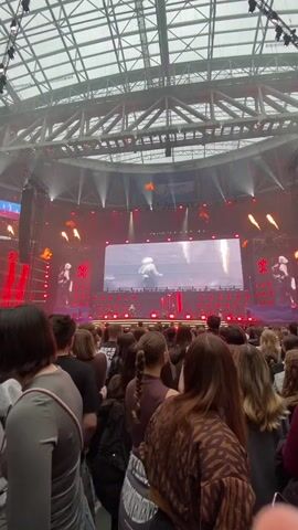 The video shows a band performing on a stage in front of a large crowd of people. The band is visible on a large screen behind the stage. There are flames shooting up from the stage. The crowd is dense and faces the stage. The event is taking place indoors with a high, arched ceiling supported by a web of metal beams. Stage lighting shines in red and yellow hues.
