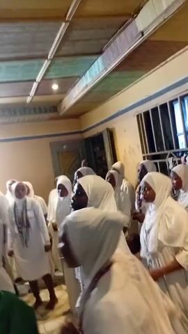 A group of people, all wearing white clothing and head coverings, are gathered in what appears to be an indoor space. They are facing each other and some are raising their hands. The room has a tiled ceiling and plain walls. There are windows with bars on one side of the room. The overall atmosphere suggests a gathering or a ceremony.