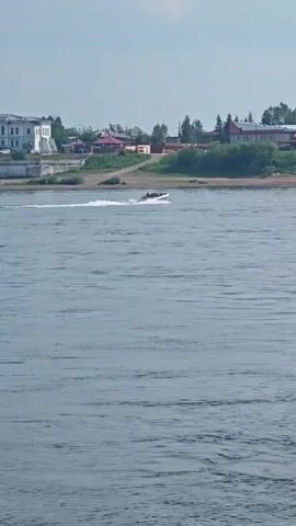 A speedboat is moving across a wide river, leaving a white wake. Several people are visible in the boat. In the background, there is a green shoreline with trees and buildings, including a white building with columns on the left and structures with blue domes on the right. The sky is partly cloudy.