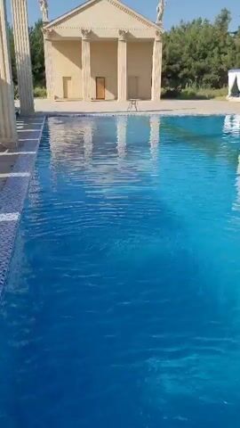 The video shows a swimming pool with clear blue water. On the far side of the pool, there is a building designed in a classical style, with columns and statues. The building appears to be an architectural replica of an ancient Greek or Roman temple. The pool is bordered by a tiled edge and a decorative mosaic pattern. The overall environment suggests a resort or a private residence with a focus on classical aesthetics. There are no people visible in the video. The water in the pool is calm, with subtle ripples indicating movement, possibly from a filter system. Trees are visible in the background, behind the classical building, suggesting an outdoor setting.