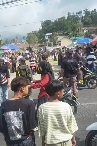 The video shows a busy outdoor scene with many people and motorcycles. The environment appears to be a street or market area, with buildings and trees visible in the background. Several people are riding motorcycles, while others are standing around or walking. Some individuals are wearing traditional Islamic clothing, such as hijabs and long dresses. There are also some vendors or stalls set up, with items displayed for sale. The overall atmosphere seems to be one of activity and gathering.