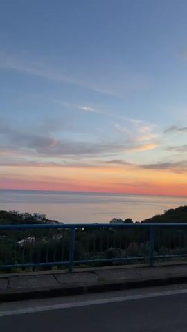 The video shows a sunset over the ocean from an elevated viewpoint, possibly a bridge or a cliffside road. A blue railing runs along the foreground, separating the viewer from a densely vegetated slope. Beyond the vegetation, the ocean stretches to the horizon, meeting a sky filled with pastel-colored clouds, predominantly shades of pink, orange, and blue. The sun itself is not directly visible, but its light is casting a warm glow across the scene. A small cluster of buildings is visible on a distant hillside. The road in the foreground has a visible white line marking a lane. There are no people or animals visible in the video. The overall atmosphere is serene and peaceful.