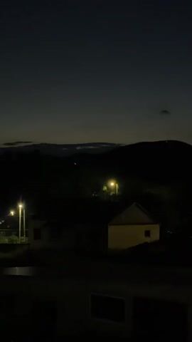 A vertical video shows a dimly lit rural landscape at dusk or dawn. Silhouetted hills form the background. In the middle ground, a few structures, possibly houses, are visible. One building has a light on inside, casting a yellow glow. Streetlights illuminate the scene with their warm light. The sky is a deep, dark blue, with a hint of lighter tones near the horizon. A single, small, dark cloud is present in the sky. The foreground shows the edge of a dark structure, likely a building or balcony, with windows visible. The overall atmosphere is tranquil and serene.