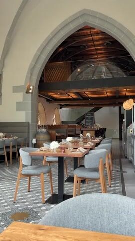 The video shows the interior of a restaurant. The restaurant is set up in a large room with a high, arched ceiling and gray stone pillars. There are rows of square tables with gray chairs, each set with a wooden plate, glassware, and a small bouquet of white flowers. In the background, there is a mezzanine level with glass railings and a dark wood beam structure. A staircase leads up to this level. There is also a wine barrel visible, positioned near a corner. The floor is tiled in a gray and white geometric pattern.
