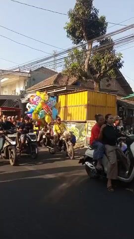 The video shows a street scene with several people on motorcycles. In the center, a motorcycle carries a large bunch of colorful balloons. People are riding on the motorcycles, and some are walking or standing on the side of the road. Buildings and trees are visible in the background, along with power lines. The lighting suggests it is daytime.