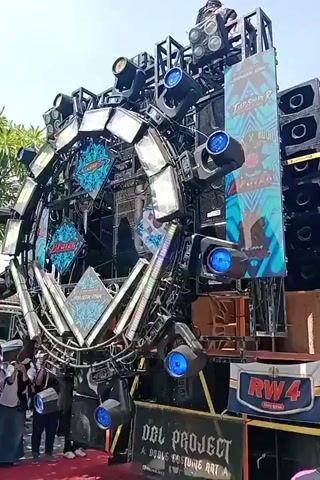 A large sound system with numerous speakers and elaborate lighting fixtures is set up outdoors. The sound system features a central circular structure adorned with lights and decorative elements, surrounded by multiple tiers of speakers. Several spotlights and colored LED lights are integrated into the structure, some flashing in sync with the music. A banner with text, including "RW4" and "DDL PROJECT," is visible on the front of the sound system. A few individuals, dressed casually and wearing headscarves, are standing in front of the sound system, some holding cameras or phones. The ground is covered with a red carpet. The scene suggests an outdoor event or performance, likely a concert or festival.