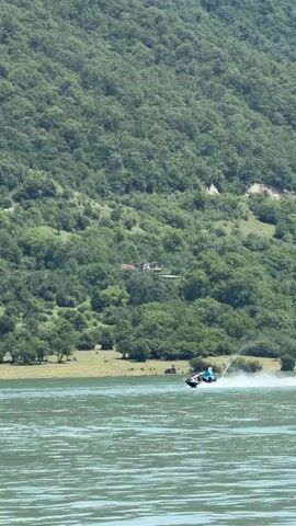 A person rides a jet ski across a body of water, leaving a wake behind them. The jet ski is black and blue and the rider is wearing a blue shirt. In the background, a lush green hillside covered in trees rises up from the water's edge. The sky is overcast.