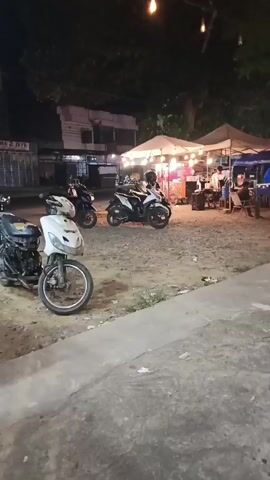 The video shows an outdoor market or food court at night. There are several food stalls set up under canopies, illuminated by string lights. Several motorcycles are parked in the foreground and middle ground. People are seen sitting at tables and interacting at the stalls. The ground appears to be gravel and dirt. The overall impression is that of a bustling night market.
