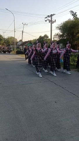 A group of young women in matching plaid dresses and pink hijabs are performing a synchronized march on an asphalt road. They are walking in formation, with their arms extended forward in a coordinated gesture. The road is lined with trees and utility poles. In the background, a person in a blue shirt appears to be directing or observing the group. The lighting suggests it is either early morning or late afternoon. A motorcycle's side mirror is visible in the foreground, indicating the perspective is from someone on a motorcycle.