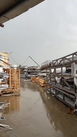 This media file captures an outdoor scene at what appears to be a storage yard or industrial area. The ground is wet and reflects the overcast sky, indicating that it is raining. Numerous stacks of materials are visible, including what seem to be wooden pallets, metal bars or pipes, and other assorted items. These are stored on wooden racks and directly on the ground. The overall impression is one of a working environment that is temporarily halted due to the weather.

The sky is a uniform, dull gray, suggesting either early morning or late afternoon, or simply a heavily overcast day. The rain is clearly visible as streaks falling from the sky, and its impact on the wet surface is evident. There are no people visible in the frame, contributing to a sense of quiet or abandonment due to the inclement weather. The setting appears to be a functional, utilitarian space rather than a residential or scenic one, with the focus being on the storage of goods. The presence of industrial-looking shelving units and the variety of materials stored suggest a business involved in manufacturing, construction, or distribution.

There is no visible text in the media file. The mood is subdued, primarily due to the weather and the absence of human activity. The rain creates a sense of dampness and a potentially cold or cool atmosphere. The location context of Struma, Bulgaria, doesn't offer specific visual cues within the image itself, but it places the scene geographically.