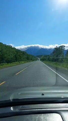 The video shows a first-person view from a car driving on a paved road. The road is surrounded by lush green trees and vegetation. In the distance, a majestic mountain range is visible, with its peaks partially obscured by white clouds. The sky is a clear, bright blue, with the sun shining prominently, creating a lens flare effect on the windshield. There are a few other vehicles on the road, including a truck that passes by. The overall scene depicts a scenic drive through a natural landscape.