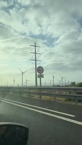 The video shows a view from inside a moving vehicle on a highway. The highway has two lanes with white dashed lines marking the lanes. A guardrail separates the highway from a flat, open area. In the background, a large wind farm with numerous wind turbines is visible. The turbines have their blades spinning. Power lines and a tall utility pole are also present. The sky is cloudy, with patches of blue sky visible. A blue and white road sign with an arrow is placed on the side of the road. A black car is seen in the left lane of the highway. The overall environment appears to be an industrial or coastal area.
