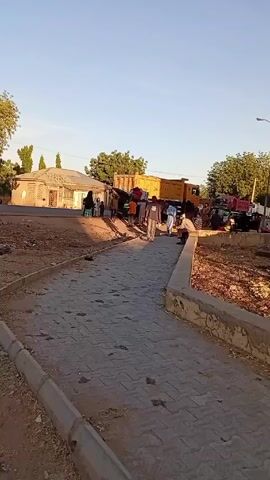 The video shows a paved sidewalk leading towards a small gathering of people. Several individuals are visible near the sidewalk and a tan-colored building with a tiled roof. A large, yellow dump truck is parked in the background. The environment includes trees, a blue sky, and areas of dry, bare ground. People are walking around. It appears to be a casual outdoor setting in a rural or suburban area.
