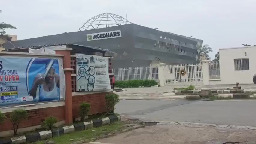 The video shows a street-level view of the ACEDHARS building. The building is a modern, dark gray structure with a geodesic dome on top. In front of the building, there is a fence and some greenery. To the left of the ACEDHARS building, there is a smaller brick structure with a sign advertising "ADMISSION FORM FOR 2025/2026 ACADEMIC SESSION ON SALE". The sign also features images related to education and swimming. The weather appears to be overcast. The camera pans slightly to the right, revealing a white building and a gate.