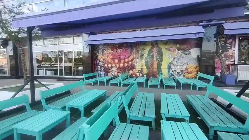 The video shows an outdoor dining area with turquoise picnic tables and benches. The seating area is covered by a purple awning and located along a waterfront promenade. The building behind the dining area has a mural depicting various images, including the Virgin Mary, a chihuahua, and sugar skulls. Across the promenade, there is a river with a cityscape in the background, featuring cranes and skyscrapers. The sky is blue with scattered clouds. The scene is empty, with no people visible.
