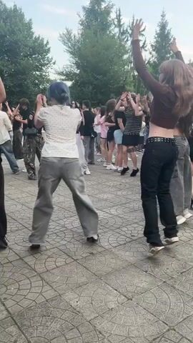 The video shows a group of teenagers dancing in an outdoor area with a paved surface. The area is surrounded by trees and a building. Some of the teenagers are dressed in casual clothing, while others are in more fashionable attire. The dancing appears to be energetic and informal, with some individuals dancing in pairs and others in a group. There is a sense of celebration and fun in the air. The video is shot from a handheld perspective, and the camera moves around to capture different angles of the dancing. The lighting suggests it is daytime, possibly late afternoon or early evening, with a bright sky and shadows.