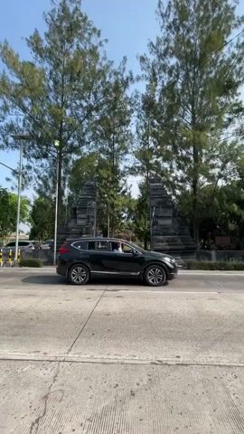 A black SUV is parked on the right side of a paved road. In the background, two stone structures resembling ancient gates stand beneath tall trees. A person rides a scooter from left to right across the road in front of the gates. The sky is clear and blue.