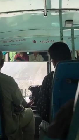 The video shows the interior of a bus, viewed from a passenger's perspective. In the foreground, a person's legs are visible, wearing light-colored pants. To their right, a person in a patterned shirt is seated, with their hand resting on their lap. Behind them, the back of a blue seat is visible. Above the windshield, a sign is visible with red text reading "No smoking". Through the windshield, a mountainous landscape is seen, with a road winding through it. Several vehicles, including cars and a truck, are visible on the road. In the upper portion of the windshield, the faces of passengers are visible, looking forward. The lighting suggests it is daytime.