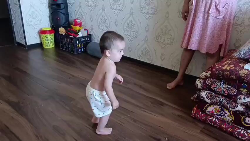 A toddler wearing white patterned briefs stands on a wooden floor in front of a patterned wallpaper. To the left, a black speaker sits near a collection of toys and a yellow object. A person in a pink dress is partially visible to the right, with their legs and feet bare. Next to them are stacked colorful patterned blankets. The toddler looks towards the right and then falls to the floor.