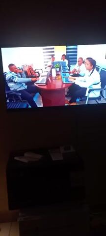 The video shows a dimly lit room with a television mounted on the wall. The television displays a scene of a meeting in a bright, modern office. There are six individuals around a circular, dark red table. Each person has a laptop in front of them. Most are wearing white coats or shirts, suggesting a professional or medical setting. One man, seated to the left, is wearing a patterned sweater and appears to be resting his chin on his hand. Below the television, there's a dark piece of furniture. Two white slippers are visible on top of it, along with some papers or a book.