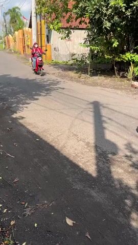 Two people are riding on a red electric scooter. The person in front is wearing a pink hijab, blue jeans, and a red jacket, and appears to be an adult. The person behind is a child wearing a dark shirt and backpack, sitting behind the adult. They are riding on a paved road with trees and buildings in the background. The sun is casting shadows on the road. The scooter is moving away from the camera.