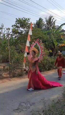 A young woman walks along a paved road, dressed in an elaborate costume. The costume features a bright pink skirt with a long train, a pink and orange bodice with gold detailing, and an ornate headpiece adorned with white, pink, and red decorations. She wears white Crocs. She is walking from right to left in the frame. To her right, slightly behind her, a man in a red tunic and head covering walks in the same direction. The environment is outdoors, with trees, bushes, and houses visible in the background. The sky is blue and sunny. There are red and white flags visible on a pole to the right of the woman.