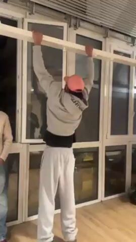 A man in a gray sweatsuit and pink hat is doing pull-ups on a wooden bar set up against a wall of windows. The man is facing away from the camera. He performs several repetitions with visible effort. Another person, wearing a light-colored sweater and jeans, stands watching from the side. The room has a wooden floor and the light is somewhat dim.  A white backdrop on a stand is visible in the background.