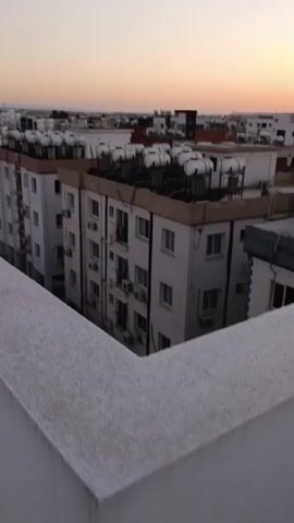 A video shows an aerial view of a city as the sun sets. Buildings with white exteriors and brown accents are visible, with rows of air conditioning units mounted on their sides. Rooftops are adorned with numerous white cylindrical water heaters. The sky displays a gradient of colors, transitioning from pale yellow near the horizon to a soft orange and pink. The scene appears to be captured from a high vantage point, possibly a rooftop or balcony, overlooking a residential or urban area. No people or significant actions are depicted.