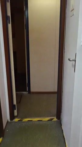 The video shows an interior room with a door ajar, revealing a room with several men. The men are wearing work uniforms in red and orange and are sitting around a table. One man is smoking and drinking from a glass. Another man is looking at a phone. The room has a small window, a clock, and some small cabinets.