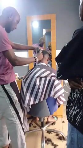 The video shows a barber cutting a client's hair in a barbershop. The barber, wearing a pink t-shirt and grey and white striped pants, is using clippers to cut the hair of a person sitting in a barber chair. The client is wearing a black and white striped cape. Another person, wearing a black jacket, stands to the right of the frame, observing the haircut. The barbershop has a mirror on the back wall, reflecting the scene, and a wooden cabinet. Hair clippings are scattered on the floor around the barber chair.
