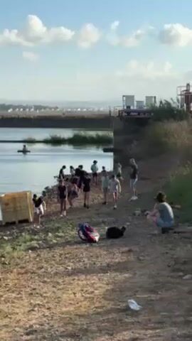 The video shows a group of people, mostly younger individuals, gathered by the edge of a body of water. They appear to be walking or milling around near the shoreline. To the right, an adult male stands observing, while a woman sits on the ground. Several bags and other personal items are scattered on the bank. In the background, across the water, are structures resembling buildings or facilities. The sky is partly cloudy, and the overall lighting suggests a sunny day. There are some industrial-looking fixtures visible along the water. The terrain is rocky and grassy.