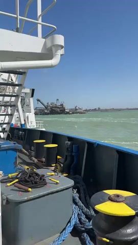 The video, filmed from the deck of a moving vessel, captures an industrial port facility under a clear blue sky. In the foreground on the vessel, white railings, a blue-painted edge, and a grey storage box with yellow and black bitts and coiled ropes are visible. The water, a greenish-turquoise, is choppy with visible waves, indicating movement. On the distant shore, a large industrial complex dominates the scene. It features multiple gantry cranes, conveyor systems, and various buildings, likely for bulk cargo handling. A sign displaying the text "FAM" is identifiable on one of the larger structures. The sunny weather and bright light are evident.