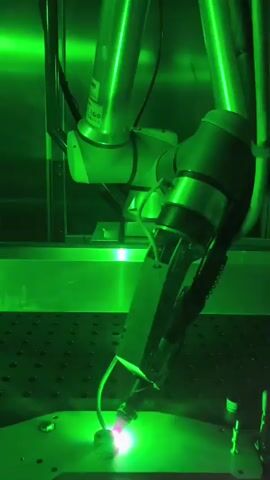 A robotic arm with a green laser welding tool is actively welding a metal object on a black surface with a grid pattern. The laser creates a bright purple and pink glow at the point of contact. Green light illuminates the scene, reflecting off the robotic arm and the metal surface.