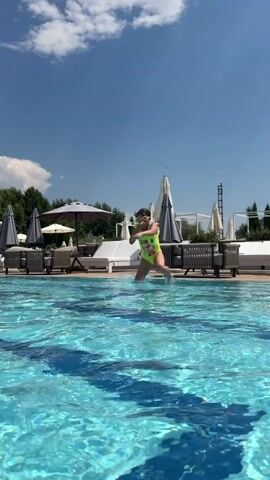 A person is swimming in a pool, with their head and arms visible above the water. The pool is outdoors, with lounge chairs and umbrellas set up around it. The sky is blue with some clouds, and the sun appears to be shining, suggesting a warm, sunny day. Trees and foliage are visible in the background, along with structures that look like part of a resort or hotel. The water is clear and blue, with sunlight reflecting off its surface. The overall scene conveys a sense of relaxation and enjoyment of a summer day by the pool.