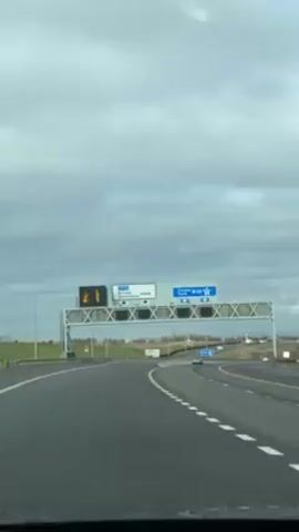 The video is shot from the interior of a car driving on a highway. Above the highway, a large overhead sign indicates that driving straight leads to Dundee and Perth on the M90.  To the left of the sign, another sign indicates directions to the M9 and South Queensferry A904. The sky is overcast. Other vehicles can be seen in the distance on the highway.