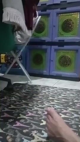 The video shows a person's foot in the foreground, lying on a patterned rug. In the background, there is a blue storage cabinet with green doors that have circular designs. To the right of the cabinet are several bags and a pink laundry basket. The scene appears to be indoors.