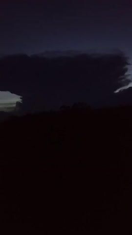 The video presents a static, silhouetted landscape at twilight. A dark, heavy cloud formation stretches horizontally across the upper portion of the frame. The silhouette of trees is visible at the midground. The foreground is entirely black, indicating the low light level. No people, objects, or actions are discernible beyond the environmental elements described.