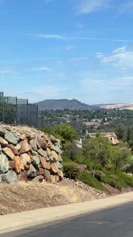 The video pans across a suburban landscape on a clear, sunny day. In the foreground, a retaining wall made of large, uneven rocks is visible on the left, with dry grass and dirt below. A paved road curves into the frame from the bottom right. Beyond the wall and road, a hillside slopes downwards, covered in green bushes and trees. Several houses with tile roofs and some with solar panels are nestled among the foliage. The middle ground features a rolling landscape dotted with more homes and trees. In the distance, hazy hills with dry, golden grass rise against a bright blue sky with sparse white clouds. The overall impression is of a warm, sunny day in a developed, hilly area.