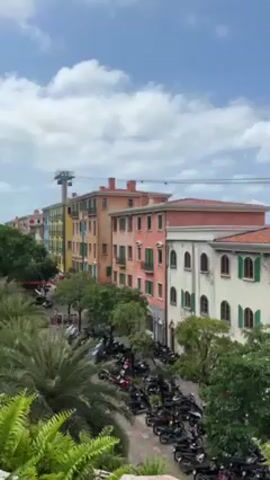 The camera pans from left to right, showcasing a scenic view. Initially, a line of colorful buildings resembling Italian architecture is visible. A street lined with trees and parked motorcycles lies below. As the camera pans, it reveals a cable car system with cars suspended on wires above the buildings. The cable cars travel towards the ocean, where several boats are visible on the water against a backdrop of a partly cloudy blue sky. Palm trees and greenery surround the buildings.