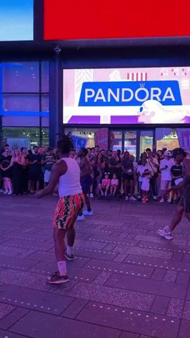 A Black male street performer is dancing in front of a crowd in an urban plaza. He is shirtless, wearing black shorts and white socks with athletic shoes. The crowd consists of approximately thirty people of various ages and ethnicities, some of whom are holding cell phones and recording. Behind the performer and crowd, there is a building with large electronic billboards displaying advertisements for Pandora and Sephora. The ground is paved with wooden planks. A young Black male is seated on a chair to the left of the frame, playing a musical instrument. Orange traffic cones are visible on the ground. The overall lighting is dim and purple.