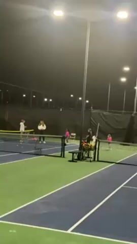 The video shows a tennis court at night, illuminated by bright overhead lights. There are two tennis courts visible, each with blue and green surfaces separated by a net. On the left court, two individuals, one wearing white and the other wearing black, are practicing tennis. A woman wearing a black top is coaching a group of children on the right tennis court. Two young children are near her; both are wearing pink outfits. The setting appears to be an indoor tennis facility, with dark screening material forming the backdrop. A ball hopper and a small bench are placed near the coach. Several tennis balls are scattered on the courts.