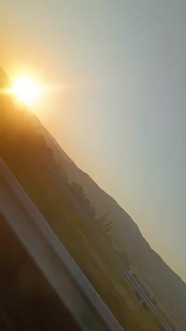 The video captures a sunset over a mountainous landscape, viewed from a moving vehicle. The sun is positioned on the left side of the frame, casting a bright orange glow and lens flare. A highway or road runs diagonally across the foreground, with its guardrail visible. The mountains are in the background, with trees and vegetation dotting their slopes. The sky is a gradient of pale blue and orange. The camera appears to be tilted, giving the impression of a diagonal perspective. The video starts with a relatively stable shot, then shifts to a more blurred and dynamic view, suggesting an increase in speed or camera movement.