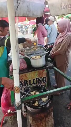 A street vendor is frying food in a large wok, with several customers waiting nearby. The vendor is wearing a green apron and glasses, and is using tongs to flip food in the hot oil. A woman in a pink hijab is standing to the right of the vendor, looking at the food. In the background, there are other people and a busy street with motorbikes. The vendor's cart has a sign that says "ARLUNG NASI". The overall scene is one of a bustling street food stall.