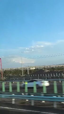 The video shows a view from a moving vehicle looking out at a highway with traffic. The sky is blue with some clouds. On the highway, there are cars and trucks, including a white sedan, a blue truck, a purple tank truck, and a white truck pulling a tank trailer. There are also what appear to be wind turbines and agricultural fields in the background. The perspective suggests the video is taken from the passenger seat of a car.