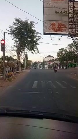 The video shows a view from inside a vehicle looking out at a road intersection. A traffic light is visible on the left, showing red. A pedestrian crossing is marked on the road. A tree stands to the left of the road, and a large, weathered billboard is visible overhead. In the distance, buildings and more trees line the road. A motorcycle is seen turning at the intersection. The sky is overcast.