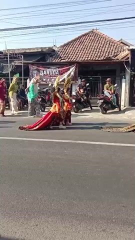 The video shows a procession of people walking on a paved road, likely during daylight hours as indicated by the clear sky and shadows. Several individuals are dressed in what appears to be traditional or ceremonial attire. One prominent figure in the foreground wears a vibrant red and gold dress with a long train, and another individual, possibly a child, is adorned in a pink costume with a large feather headdress and a matching train. Other participants are seen in more casual clothing, some wearing headscarves. They are walking from left to right across the frame. In the background, there are buildings with tiled roofs, a banner that reads "GARUDA," and motorcycles parked or being ridden. The overall atmosphere suggests a cultural event or celebration taking place in a semi-urban or village setting.