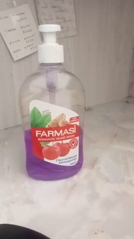 This media file shows a bottle of Farmasi Romantic Hand Wash with Cherry & Almond & Pomegranate scent. The bottle is clear plastic with a white pump dispenser and a label featuring images of cherries, almonds, and green leaves. The hand wash inside is a vibrant purple color.

The bottle is placed on a light-colored, possibly marble or laminate surface. To the left of the bottle, there are several pieces of paper with handwritten text, appearing to be notes or receipts. Some of the visible text includes names like "MUGAMBI," "TABITHA," "NKURI," "MWIRIGI," and numbers that might be phone numbers or identification codes. One note mentions "Kiemu/Igembe East ward, Kenya," providing a geographical context for the location.

The lighting suggests it is daytime, though no specific time can be determined. The overall scene appears to be an indoor setting, likely a desk or shelf, given the presence of the notes. There are no people visible in the frame. The focus is on the hand wash bottle and its immediate surroundings. The image is captured from a slightly elevated angle, and the bottle is oriented horizontally.