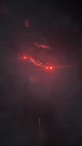 A slow-motion video captures a single red flare ascending into a dark, smoky sky. The flare ignites, emitting bright red light and sparks that trail behind it. The smoke creates a hazy, atmospheric effect around the ascending flare.
