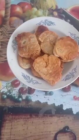 The video displays a close-up shot of a white plate filled with several golden-brown pancakes. The pancakes are piled on top of each other and appear to be fluffy and well-cooked. The plate has a decorative pattern around its rim, featuring blue leaves and vines. The plate is placed on a table covered with a colorful tablecloth that has a fruit design. In the background, there are images of various fruits, including apples, grapes, and watermelon. Below the plate, a white lace doily is visible, and the table surface beneath it has a wood grain pattern. The lighting is natural and appears to be coming from above.